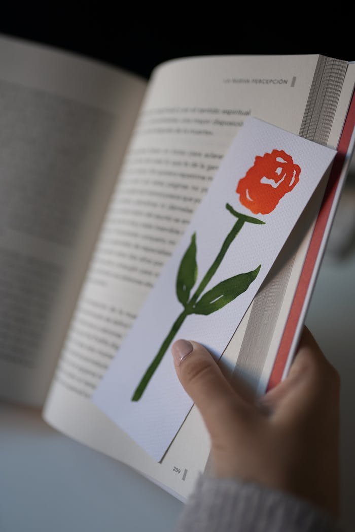 Close-up of hand holding a painted rose bookmark in an open book, perfect for reading themes.